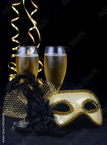 New Year's Eve themed image of gold and silver carnival mask decorated with black flower and feathers with gold ribbons on soft, dark cloth. Two glasses of champagne. Copy space.