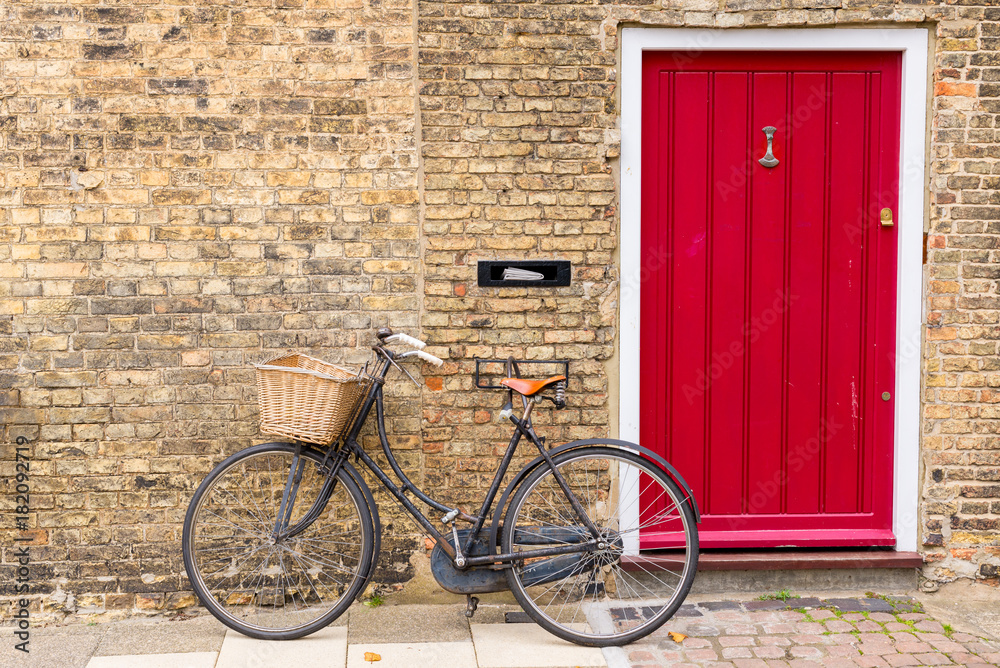 Vintage bicycle with retro wicker basket parked in front of a house brick wall next to a bright red door