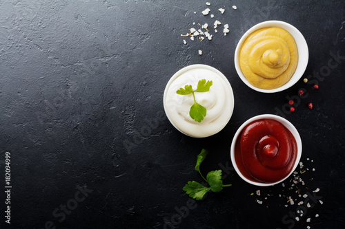 Set of three sauces - mayonnaise, mustard and ketchup in white ceramic bowls on black stone or concrete background. Selective focus. Top view.