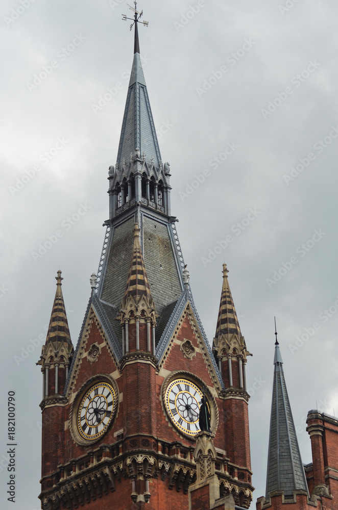 A gothic clock tower against a stormy sky. It is made of red bricks, with a grey spire. Two clock faces are visible.