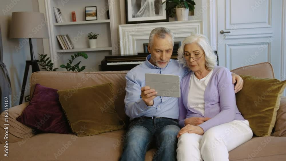 Cute grandparents using tablet while sitting on the sofa at home in the stylish living room. Side view. Cute grandparents using tablet while sitting on the sofa at home in the stylish living room. Side view.