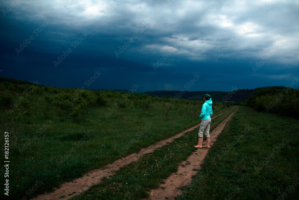 female stands on the road which extends along the green valley with bushes and trees. Rock on the horrizon, blue and grey stormy clouds female stands on the road which extends along the green valley with bushes and trees. Rock on the horrizon, blue and grey stormy clouds