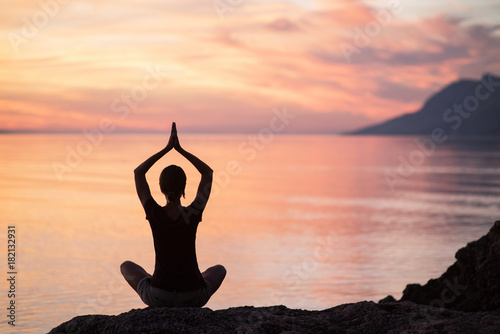 Silhouette of a young woman practicing yoga near the sea at sunset. Harmony and meditation concept. Healthy lifestyle