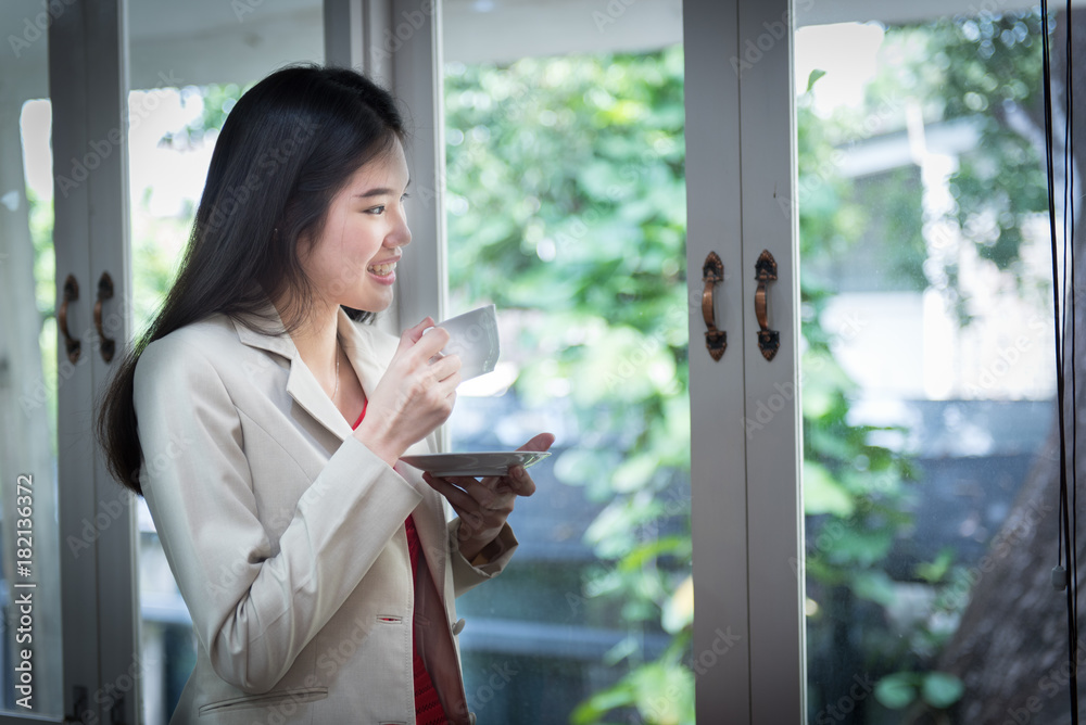 Smile of Beautiful Asian Businesswoman stand and holding coffee cup near the window. in office. business successful concept. Smile of Beautiful Asian Businesswoman stand and holding coffee cup near the window. in office. business successful concept.