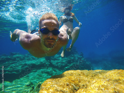 Underwater close photo of a young playful love couple swimming in the turquoise exotic sea for summer vacation.