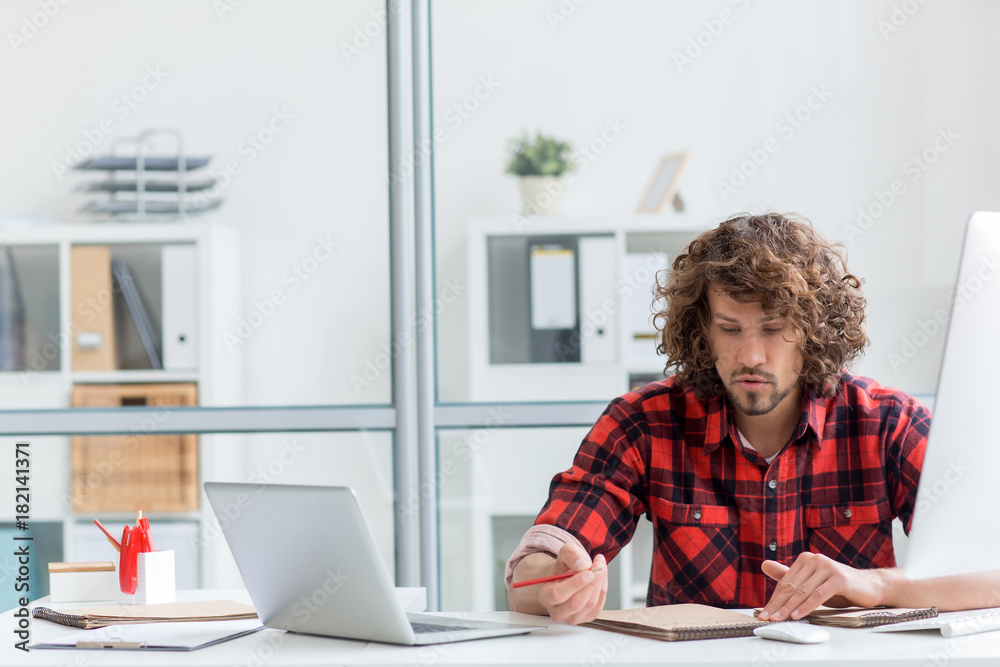 Young manager or designer with pencil making sketch in notepad by his workplace Young manager or designer with pencil making sketch in notepad by his workplace