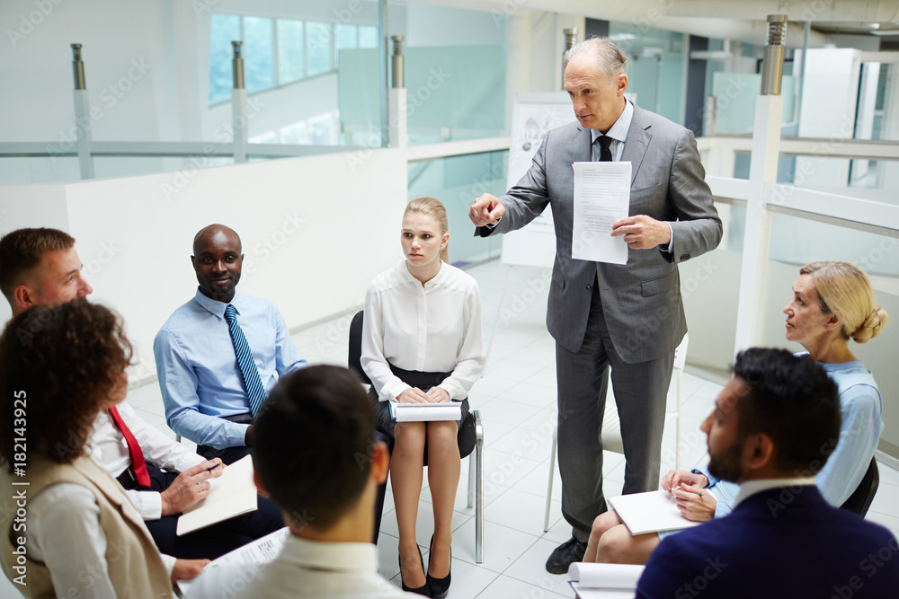 Mature financial expert with paper pointing at one of managers during explanation of data Mature financial expert with paper pointing at one of managers during explanation of data