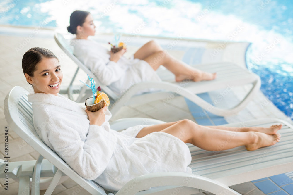 Happy young woman with tropical drink looking at camera at spa resort with her relaxing friend on background