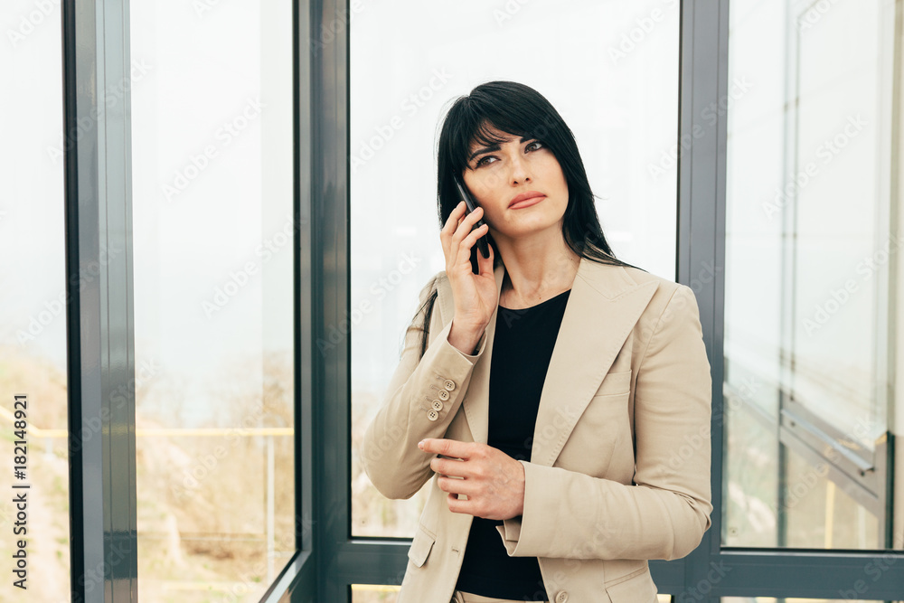a beautiful business girl in a beige strict suit are standing in a room with picturesque views and tilts her head to the left and holds a phone a beautiful business girl in a beige strict suit are standing in a room with picturesque views and tilts her head to the left and holds a phone