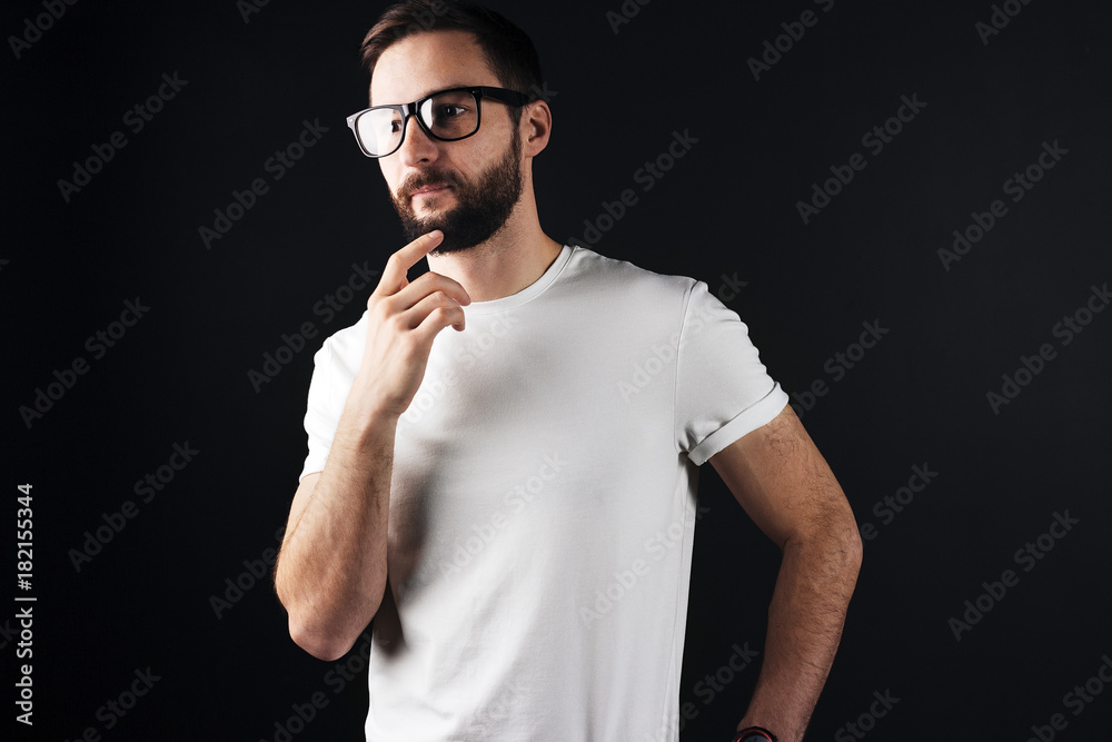 Handsome pensive bearded man in clear glasses wearing white blank tshirt in dark background. Empty t-shirt on slim body Handsome pensive bearded man in clear glasses wearing white blank tshirt in dark background. Empty t-shirt on slim body