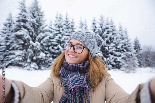 Young woman takes selfie on a background of snow-covered winter forest. Snowy weather. Woman taking photo on phone. Girl smiling at the camera. Winter holidays.