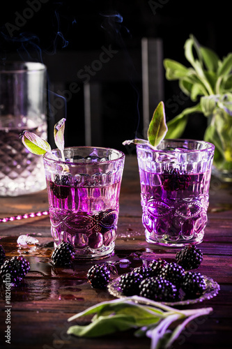 Two glasses with violet cocktails and blackberries on a wooden table with mysterious smoke
