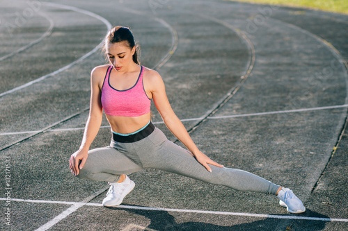 Gorgeous female runner in pink top and gray pants stretching legs before workout - outdoor shot. Teenager healthy, sport and millennials people concept.