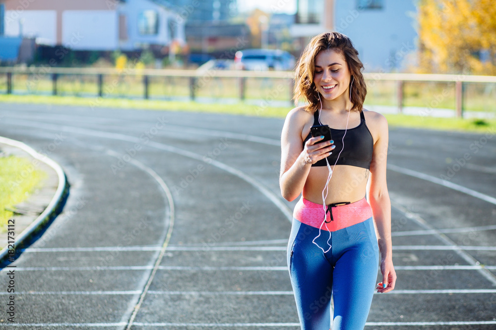 Smiling female jogger looking confident. Young fitness woman listening music with headphones after training outdoors at stadium track. Girl runner listen music in earphones from smartphone.