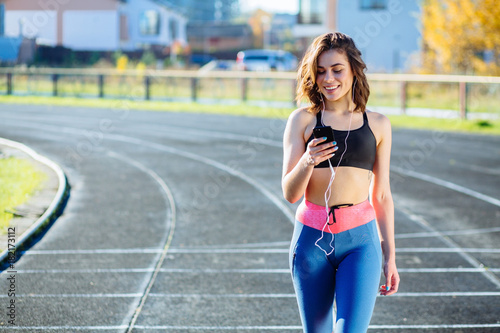 Smiling female jogger looking confident. Young fitness woman listening music with headphones after training outdoors at stadium track. Girl runner listen music in earphones from smartphone. © Iryna