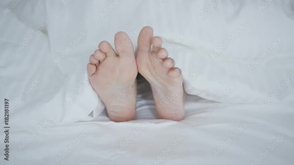 Crop shot of woman lying under cozy white blanket in bed sticking out feet while sleeping and relaxing. Crop shot of woman lying under cozy white blanket in bed sticking out feet while sleeping and relaxing.