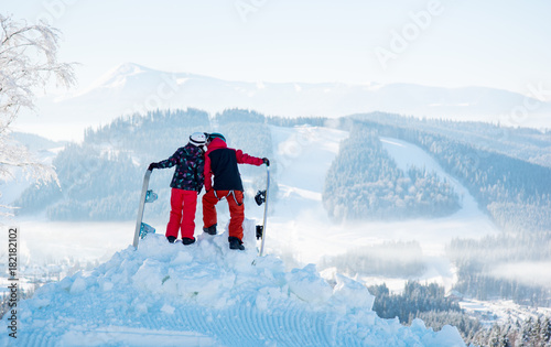 Pair of snowboarders kissing on top of the mountain against a background of snow-covered forests and hills at winter ski resort. Back view