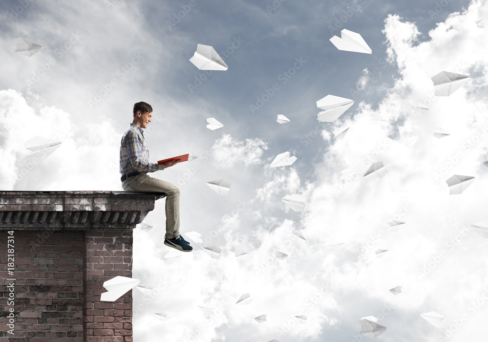 Handsome student guy on roof edge reading book and paper planes fly around Handsome student guy on roof edge reading book and paper planes fly around