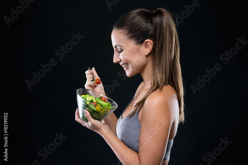 Beautiful young woman eating salad over black background.