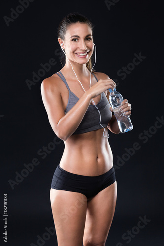 Beautiful young woman drinking water after doing exercise over black background.