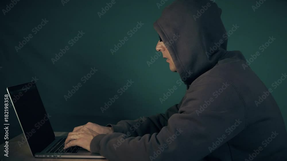 Programmer in hood and glasees typing on keyboard of laptop computer. Hacker write memo. Man got it gestures by hand. Low key side view hacker sit at table at laptop. Programmer in hood and glasees typing on keyboard of laptop computer. Hacker write memo. Man got it gestures by hand. Low key side view hacker sit at table at laptop.