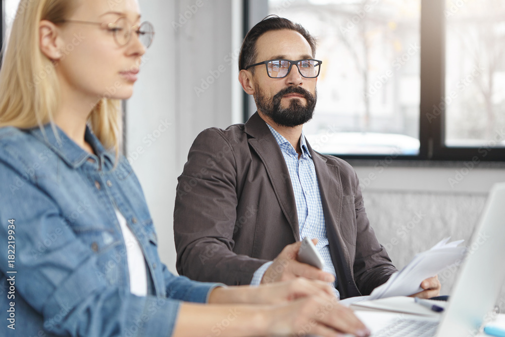 Middle aged bearded experienced male entreprsing director organizes working atmosphere, controls and motivates his assistant to finish report, has serious look in notebook, checks information Middle aged bearded experienced male entreprsing director organizes working atmosphere, controls and motivates his assistant to finish report, has serious look in notebook, checks information