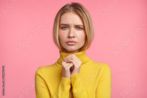 Faithful light haired female keeps palms together, hopes for better, has sad unhappy expression, isolated over pink background. Beautiful young woman asks God for health. People and wish concept