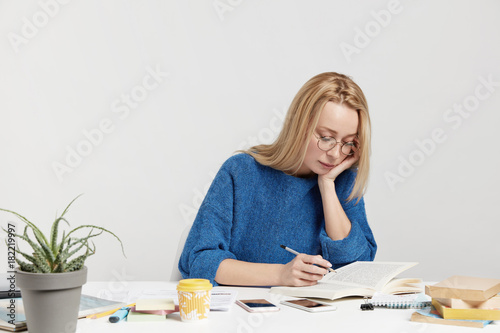 Horizontal portrait of busy female student focused in book, underlines necessary information for future report, surrounded with modern electronic gadgets and coffee. Youth and studying concept