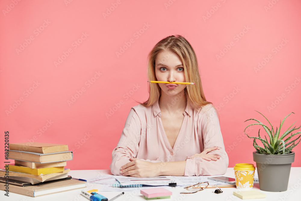 Φωτογραφία Horizontal portrait of funny female student keeps pencil on mouth, foolishes after long prepareation to pass course paper, surrounded with many books, isolated over pink background