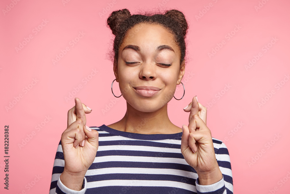 Isolated shot of attractive coquette female model with dark skin closes eyes and crosses fingers in anticipation, believes for good luck, isolated over pink studio background. Woman gestures indoors - obrazy, fototapety, plakaty 