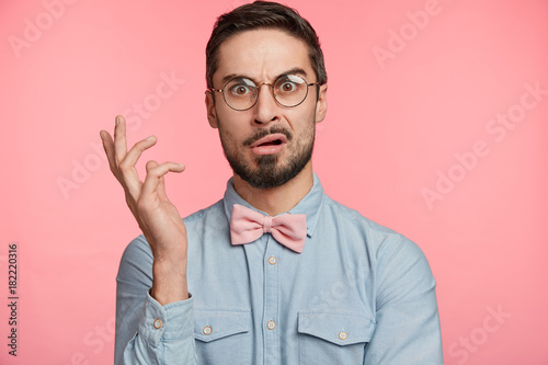 Portrait of dissatisdfied man wears round spectacles, has discontet, indigant look, gestures hand, being surprised with something, poses against pink studio background. Facial expression concept