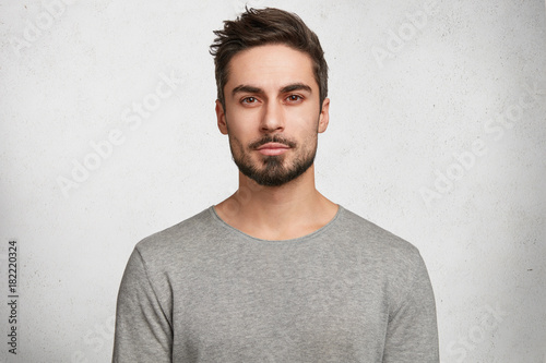 Isolated shot of young handsome male with beard, mustache and trendy hairdo, wears casual grey sweater, has serious expression as listens to interlocutor, poses in studio against white background photo