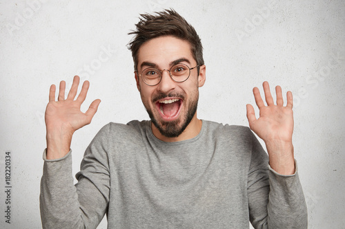 I am not guilty! Cheerful stylish fashionable young hipster guy in eyewear raises hands as shows being uninvolved, has happy look, isolated over white background. Caucasian male gestures in studio photo