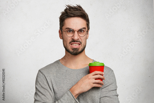 Annoyed dissatisfied young fashionable male with trendy hairdo, holds takeaway coffee, doesn`t like film which he watches, feels disgust from horrified scene, dressed in casual grey sweater. photo