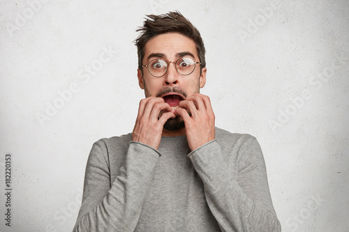 Indoor shot of horrified frightened hipster guy stares through spectacles, gestures actively and nervously, trembles from fear, has real fobia towards something, worries and afraids of darkness.