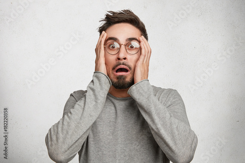 Scared European guy looks with terror in camera, can`t believe in his failure, keeps hands on head, opens mouth, has horrified expression, isolated over white wall. Frigtened young bearded male photo