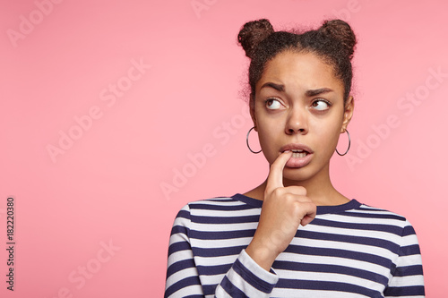 Horizontal portrait of African American female teenger has thoughtful expression, keeps finger on lips, looks aside as thinks about something important, poses against pink wall with copy space