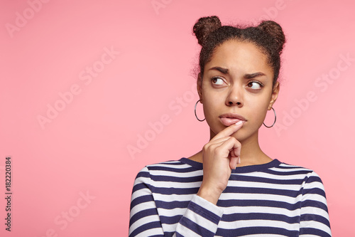 Pensive female student thinks about solving her problem, tries to predict all possible ways out, looks aside, poses against pink background with copy space for your advertisment and promotional text