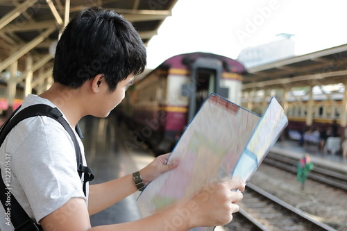 Rear view of young Asian tourist with a bag looking at map in train station with copy space background.