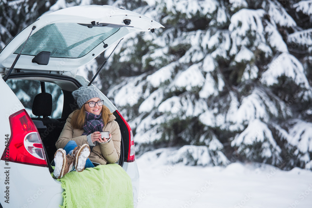Young woman in woolen hat sits in the trunk of the car and holds a cup of hot tea in her hands. Girl is smiling. Winter travel, snow-covered trees in the forest. Snowy weather.