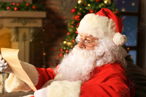 Portrait of happy Santa Claus sitting at his room at home near Christmas tree and reading Christmas letter or wish list. photo