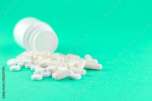 Close up white pill bottle with spilled out pills and capsules on aquamarine background with copy space. Focus on foreground, soft bokeh. Pharmacy drugstore concept