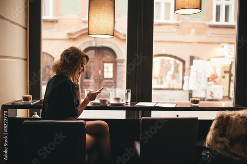 young adolescent woman in her late twenties, sitting in a bar at the window, drinking coffee and talking to a friend on a phone  photo