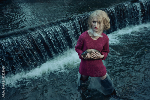 fashion portrait of model with dead fish standing in river of sewage against waterfall of dirty and polluted water