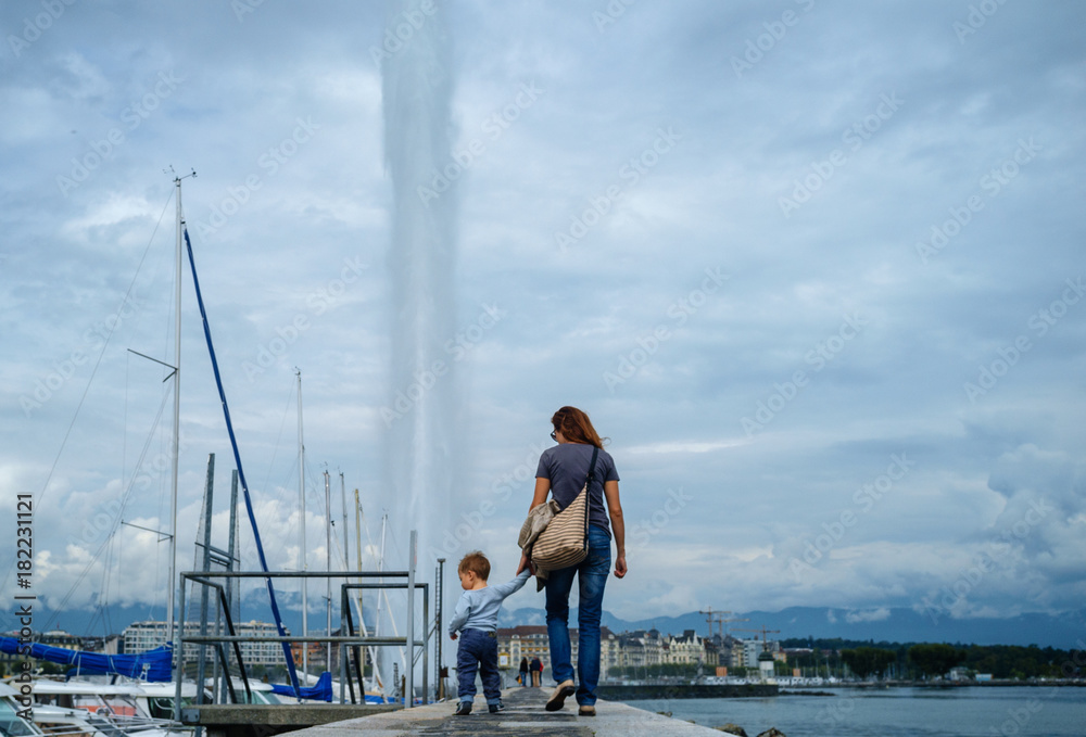 A girl with a child is holding hands on the pier and the background of a large Geneva fountain.