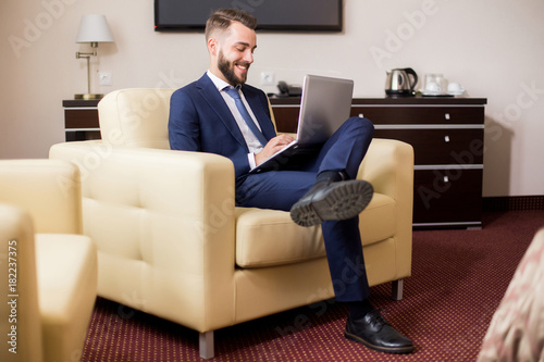 Full length portrait of handsome young businessman using laptop in hotel room and smiling while relaxing casually in armchair © Seventyfour