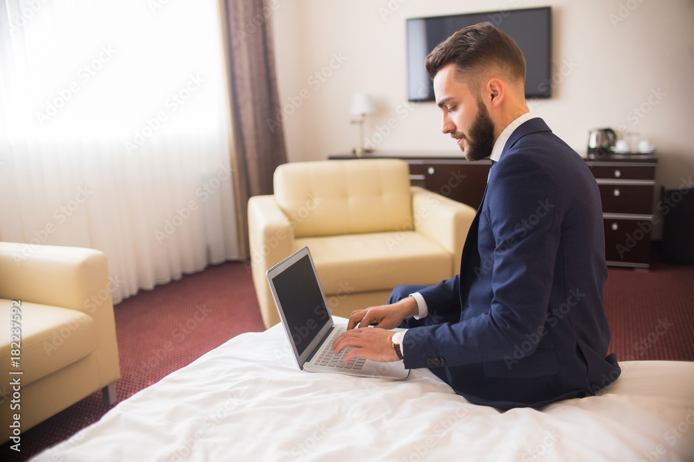 Side view portrait of handsome bearded businessman using laptop sitting on bed in comfortable hotel room during business travel, copy space Side view portrait of handsome bearded businessman using laptop sitting on bed in comfortable hotel room during business travel, copy space