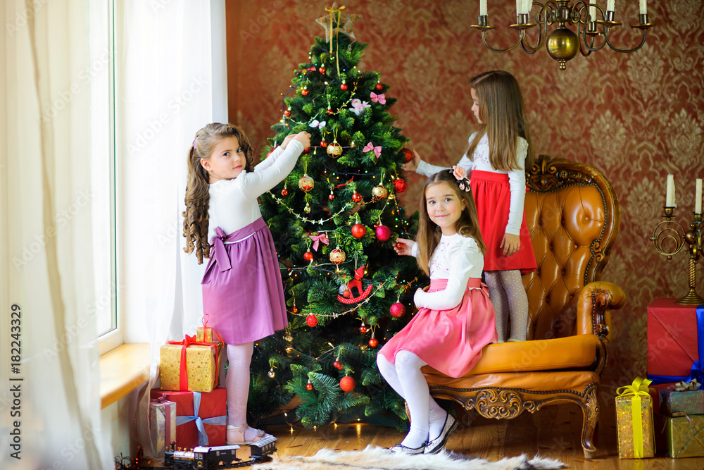 three girls in beautiful dresses decorate a festive Christmas tree, on the floor lie boxes of gifts.