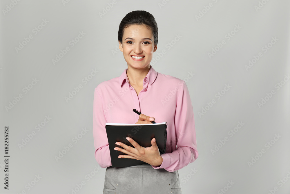 Beautiful young manager writing in notebook against grey background Beautiful young manager writing in notebook against grey background