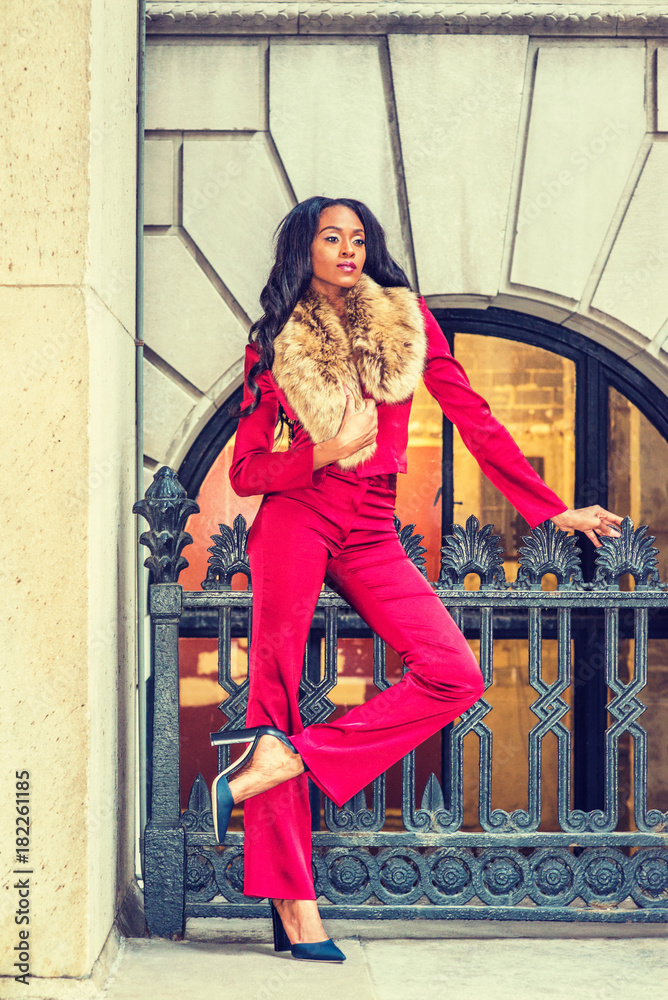 Obraz premium African American Woman Fashion in New York. Lady dressing in red slim fashionable jacket, pants, black high heel pumps shoes, faux fur collar scarf on shoulder, stands by railing. Filtered effect..